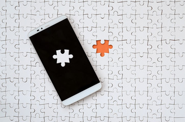 A modern big smartphone with several puzzle elements on the touch screen lies on a white jigsaw...