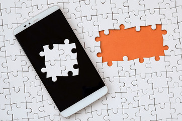 A modern big smartphone with several puzzle elements on the touch screen lies on a white jigsaw...