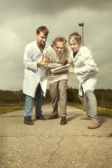 Two older retro paramedic freak hunters in medical coats catching crazy man in park