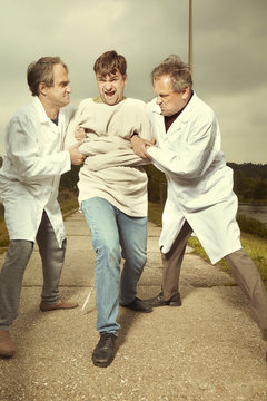 Two older retro paramedic freak hunters in medical coats catching crazy man to straitjacket