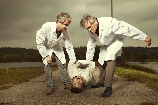 Two older retro paramedic freak hunters in medical coats catching crazy man to straitjacket