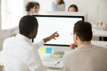 Fototapeta premium Rear view of african team leader explaining caucasian colleague corporate software usage, black manager helping coworker with computer task, mentor teaching intern, giving instructions about emails