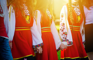 Girls in traditional Bulgarian costumes with red dresses and patterns on white shirts holding hands in the sunset. Concept of unity