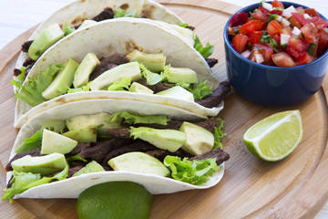 Corn tortillas with grilled beef, avocado, lime and salsa on wooden board, closeup. Side view.
