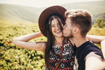 stylish hipster couple taking selfie and smiling on top of mountains in sunny summer day. beautiful man and happy woman in hat hugging and embracing on mountain. travel together