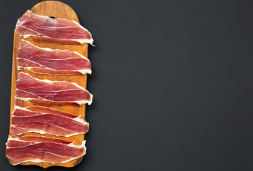 Sliced jamon Serrano or Iberico on cutting wooden board. Traditional spanish hamon on dark wooden background, top view. Copy space.
