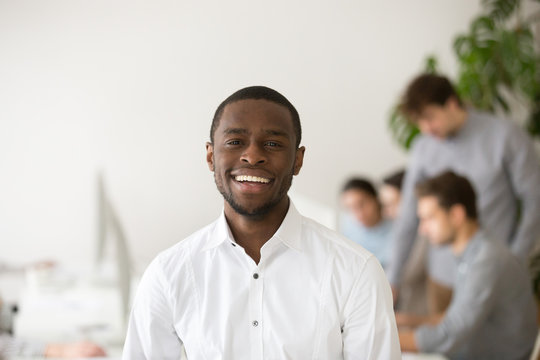 Happy african american professional smiling looking at camera with colleagues at background, friendly black business coach, successful intern, team leader member posing in office, head shot portrait