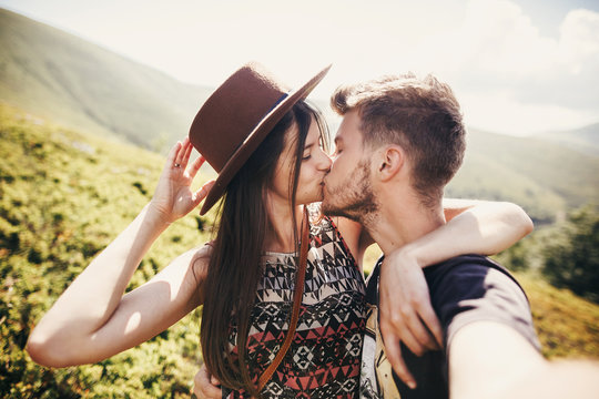 stylish hipster couple taking selfie and kissing on top of mountains in sunny summer day. beautiful man and happy woman in hat embracing on honeymoon. travel together