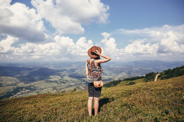 travel and wanderlust concept. stylish traveler girl in hat looking at sunny mountains and clouds sky. summer vacation. space for text. back view. amazing atmospheric moment