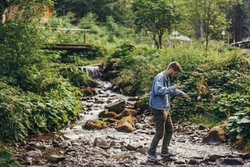 stylish traveler man in denim walking in river in sunny forest and mountains. travel and wanderlust concept. space for text. happy hipster guy traveling in woods. summer vacation