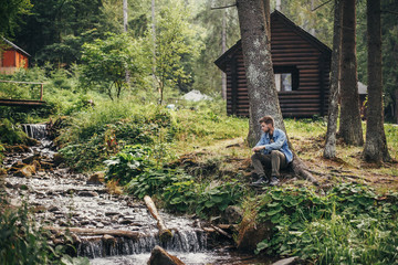 stylish traveler man sitting near cottage and river in sunny forest and mountains. travel and...