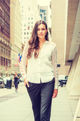 Young Eastern European Woman with long black hair, traveling in New York, wearing white, lace cut out long sleeve shirt, black pants, walking on street with high buildings in middletown of Manhattan..