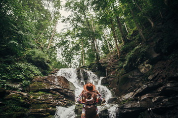 Obraz premium stylish hipster girl in hat with backpack looking at waterfall in forest in mountains. traveler woman exploring woods. travel and wanderlust concept. atmospheric moment