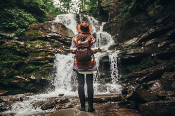 travel and wanderlust concept. stylish hipster girl in hat with backpack looking at waterfall in...