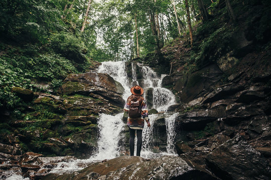 stylish hipster girl in hat with backpack looking at waterfall in forest in mountains. traveler woman exploring woods. travel and wanderlust concept. atmospheric moment