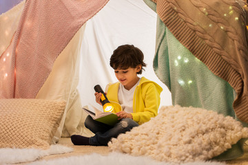 childhood, hygge and people concept - happy boy reading book with torch light in kids tent or teepee at home © Syda Productions