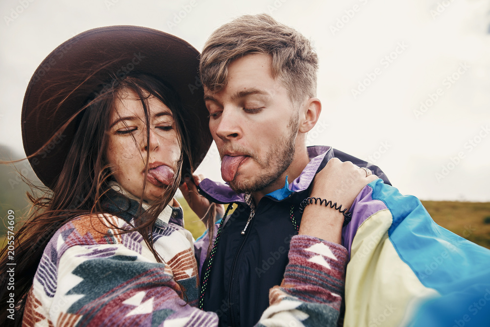 Wall mural happy hipster couple making selfie and showing colored tongues from blueberries on top of sunny mountains. family having fun and smiling. summer vacation. space for text.