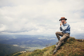 Naklejka na ściany i meble travel and wanderlust. stylish traveler man in hat sitting on top of mountains with photo camera in clouds. space for text. hipster guy traveling, taking images. amazing atmospheric moment.