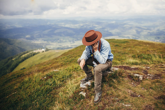 Stylish Traveler Man Sitting On Top Of  Mountains In Clouds. Space For Text. Hipster Guy Traveling, Talking On Phone. Amazing Atmospheric Moment. Travel And Wanderlust Concept