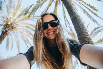 Young beautiful woman tourist or blogger makes a selfie or is reporting to its subscribers on the background of palm trees in a hot country