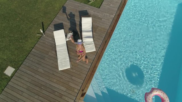 SLOW MOTION, AERIAL, POV: Angry young woman chases buzzing drone taking photos of her suntanning by her backyard pool. Annoyed blonde girl tries to crash remote controlled plane invading her privacy.