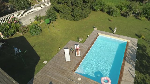 AERIAL, POV: Funny flying shot of angry Caucasian woman shouting and chasing trespassing drone around her backyard. Annoyed girl tries to crash crazy neighbor's drone buzzing above her while she tans.