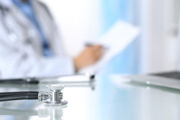 Stethoscope lying on glass desk with laptop computer at busy physician background. Medicine or pharmacy concept. Medical tools at doctor working table