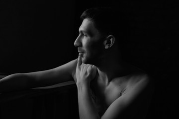 Black and white noir profile portrait of thoughtful handsome Caucasian topless male, tenderly touching his body, enjoying shape and fit, dreaming and resting in darkness. Fashion and beauty concept.