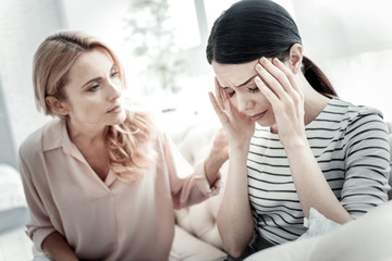 Bed feeling. Exhausted sad cute woman sitting near her friend on the sofa touching her head closing eyes.