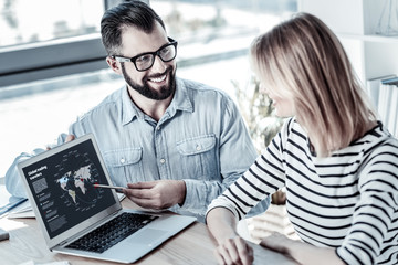 New program. Bespectacled pleasant friendly man sitting opposite his colleague smiling and demonstrating program.