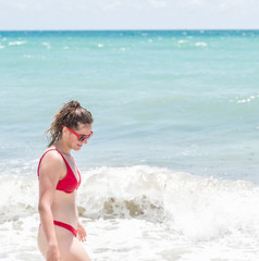 Young happy smiling woman standing in red swimsuit bikini bathing suit, sunglasses looking down at wave on Hollywood, Miami Beach in Florida side profile on sunny day, clear blue green water