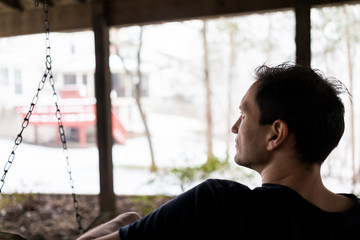 Obraz premium Closeup of young man sitting on swing under wooden deck of house on backyard in neighborhood with snow during blizzard white storm, snowflakes falling in Virginia suburb, single family home