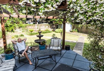 Young man lying down on patio lounge chair in outdoor spring flower garden in backyard porch of...