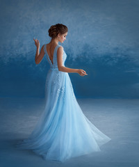 Fototapeta na wymiar A young blonde girl is spinning in a luxurious blue dress with an open back. The background is a sky-blue wall. Beautiful, elegant, high hairstyle with weaving. Evening image