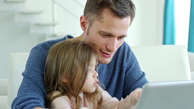 Medium shot of cute little girl sitting on laps of father working on laptop at home
