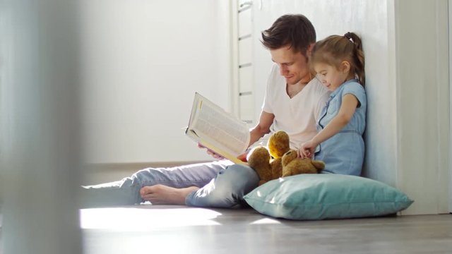 Side view of cute little girl with plush toy sitting on floor with father and listening to him reading story from book