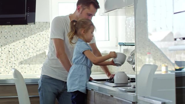 Handheld side view of father helping adorable little girl washing dishes in kitchen