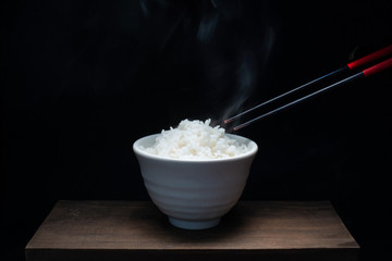 Bowl of rice and chopsticks on wood with steam, black background