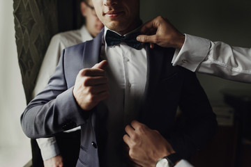 stylish groomsmen helping happy groom getting ready in the morning for wedding ceremony. luxury man in suit in room. space for text. wedding day. - Powered by Adobe