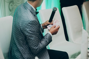 Obraz na płótnie Canvas wedding ceremony master with microphone having fun and playing games with guests. man holding and counting money at wedding reception. funny moment. space for text