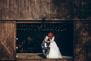 rustic wedding couple posing and hugging at background of wooden barn with retro lights, space for text. happy stylish bride and groom at wood wall in country, bohemian newlyweds