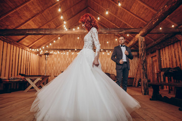 Fototapeta na wymiar stylish groom and happy bride dancing under retro bulbs lights in wooden barn. rustic wedding concept, space for text. newlyweds couple embracing, sensual romantic moment