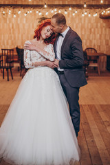 Fototapeta premium stylish groom and happy bride gently hugging under retro bulbs lights in wooden barn. rustic wedding concept, space for text. newlyweds couple embracing, sensual romantic moment