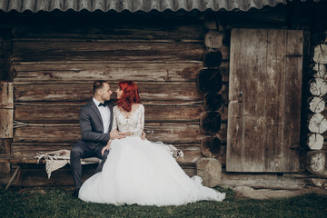 stylish groom and happy bride sitting on bench at wooden house in evening. rustic wedding  in barn...