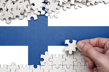 Finland flag  is depicted on a table on which the human hand folds a puzzle of white color