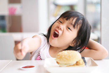 Asian children cute or kid girl delicious eating ham burger or junk food with tomato sauce or ketchup on white dish and table for breakfast or lunch with hold knife for happy fun smile at restaurant