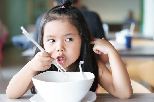 Asian Children Cute Or Kid Girl Hungry And Enjoy Eating Noodle Delicious Food In White Bowl On Wood Table And Use Chopstick For Breakfast Or Lunch At Cafe Restaurant Or Food Court