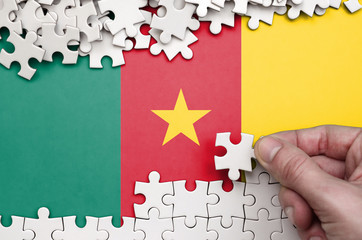 Cameroon flag  is depicted on a table on which the human hand folds a puzzle of white color