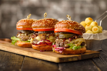 Three homemade burgers with meat, cheese, pickles, greens, tomatoes and caramelized onions on the wooden board served with fried potato balls.