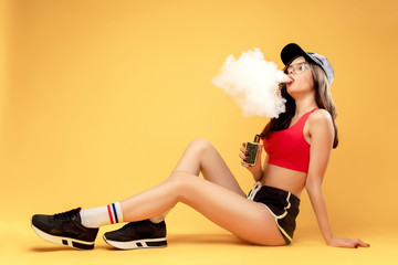 Glamour attractive woman sitting and puffing vapor on yellow background.